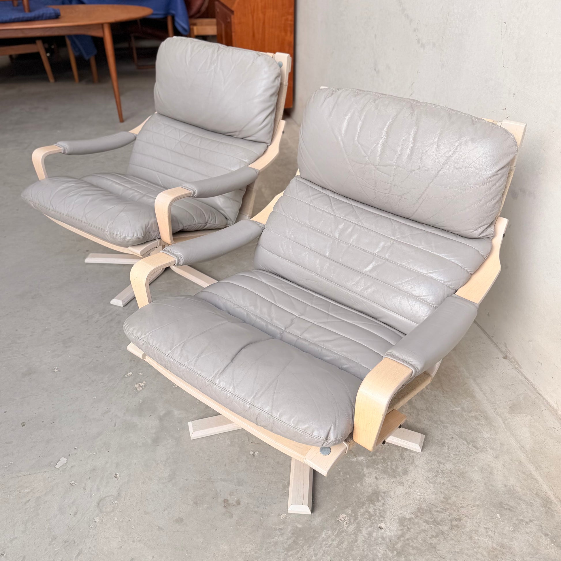 Two Tessa Ferrara Swivel Armchairs (Revive) with light wooden frames and light gray leather cushions, inspired by mid-century Australian design, sit on a concrete floor against a plain wall.