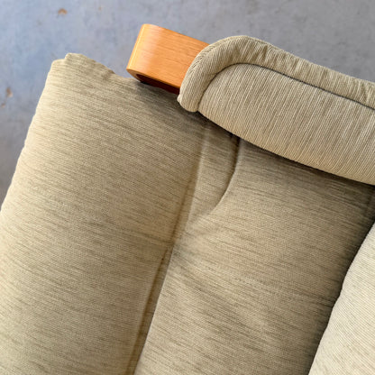 Close-up of the corner of a sage green corduroy chair with a wooden armrest, echoing mid-century style. The textured fabric and soft padding highlight the comfort of this Pair of Tessa Monaco Arm Chairs - Like New (Revive). Blurred gray background.