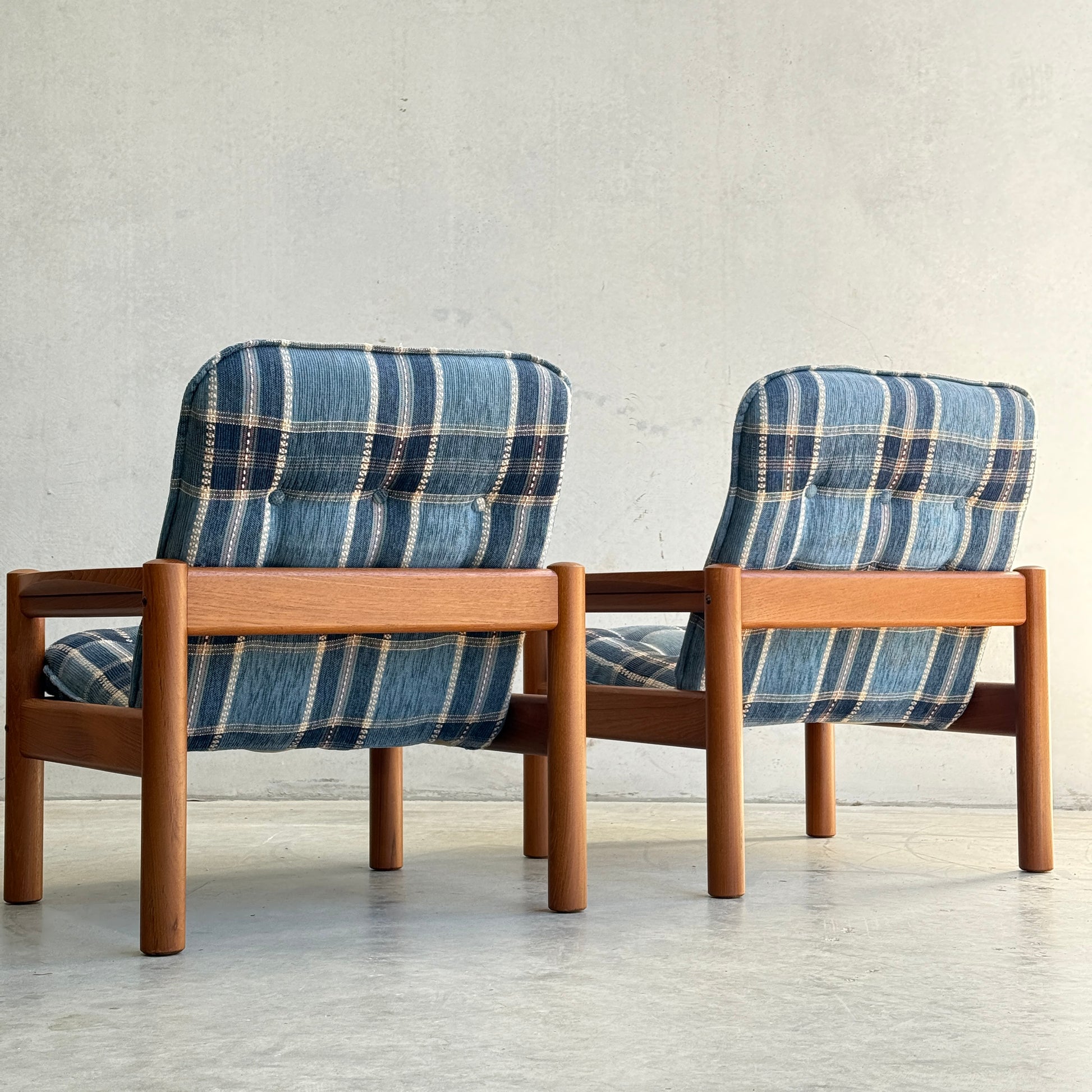 The Restored Domino Møbler Armchairs – Blue Plaid Mid-Century Set (Rediscover) features two Scandinavian seats with retro plaid cushions and solid wood frames, placed side by side against a light wall and concrete floor.