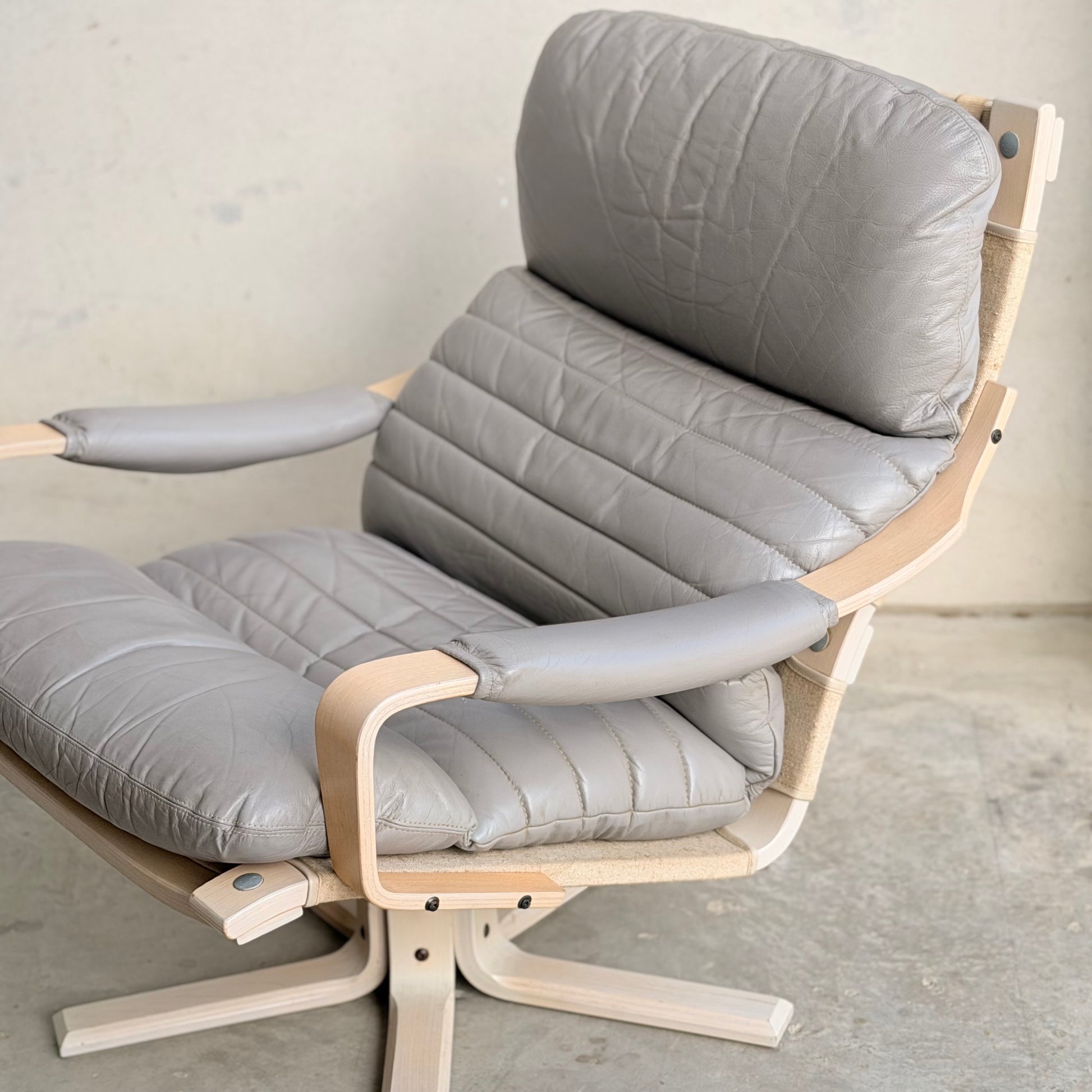 A pair of Tessa Ferrara Swivel Armchairs (Revive) with light gray leather cushions, light wood frames, armrests, and swivel bases stand out against bare concrete—an homage to mid-century Australian design.