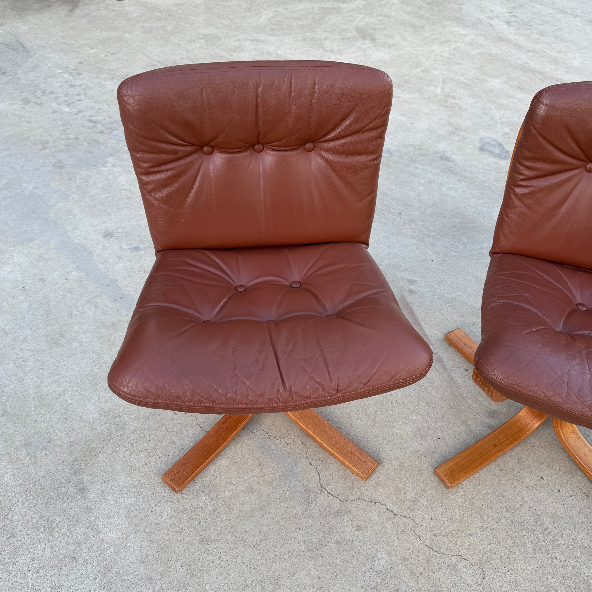 A Rare Set of 4 Vintage Joe Rufenacht Lounge Chairs in Howe Leather, featuring button-tufted upholstery and wooden crossed legs, showcases Scandinavian-inspired design on a concrete surface. Another chair from the set is partially visible on the right.