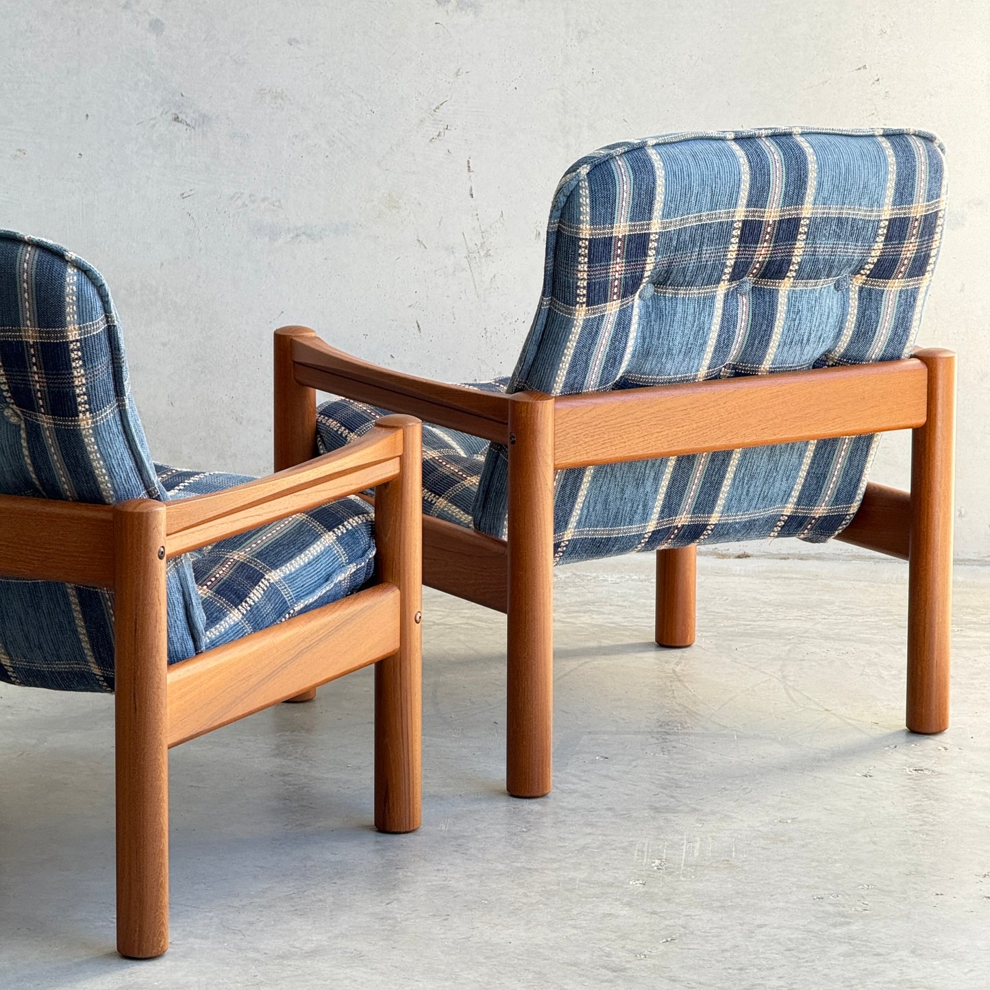 Two Restored Domino Møbler Armchairs – Blue Plaid Mid-Century Set (Rediscover) sit on a light-colored floor against a plain wall, showcasing solid wood frames, retro blue plaid cushions, and elegant Scandinavian mid-century modern design.