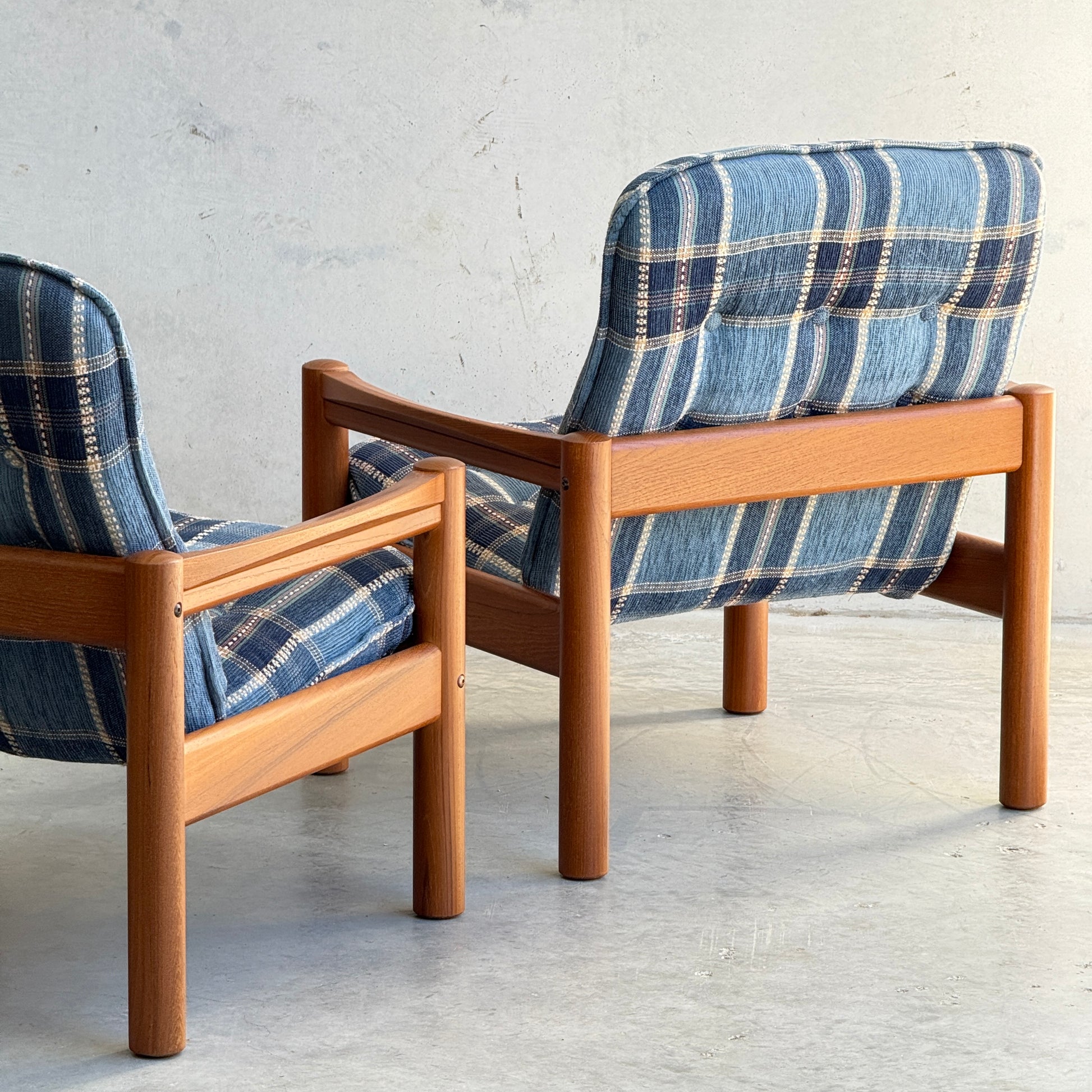 Two Restored Domino Møbler Armchairs – Blue Plaid Mid-Century Set (Rediscover) sit on a light-colored floor against a plain wall, showcasing solid wood frames, retro blue plaid cushions, and elegant Scandinavian mid-century modern design.