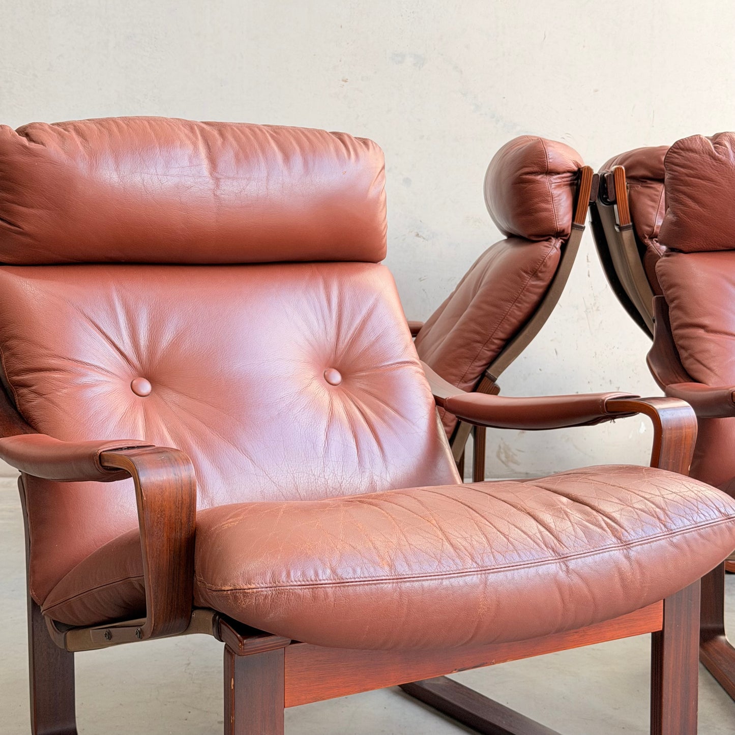 A close-up of the Tessa T8 Lounge Suite modular two-seater sofa and armchairs (Revive), featuring brown leather cushions and wooden frames, placed on a light floor against a plain, light wall.