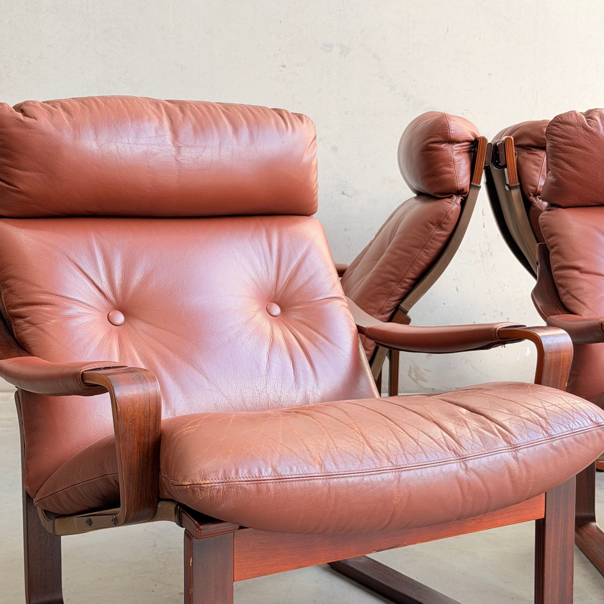 A close-up of the Tessa T8 Lounge Suite modular two-seater sofa and armchairs (Revive), featuring brown leather cushions and wooden frames, placed on a light floor against a plain, light wall.