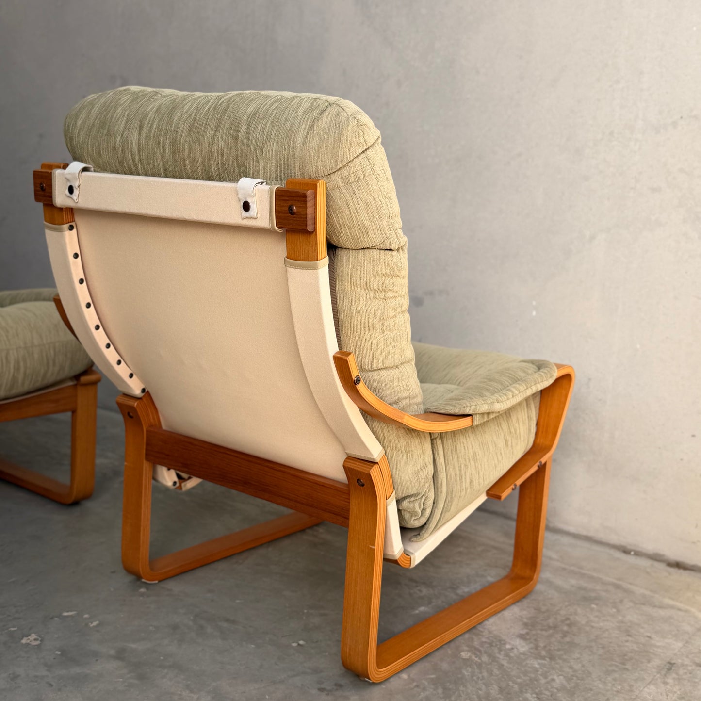 A pair of Tessa Monaco Arm Chairs in sage green corduroy, like new (Revive), with beige backs and wooden frames, are placed on a smooth concrete floor against a plain grey wall.
