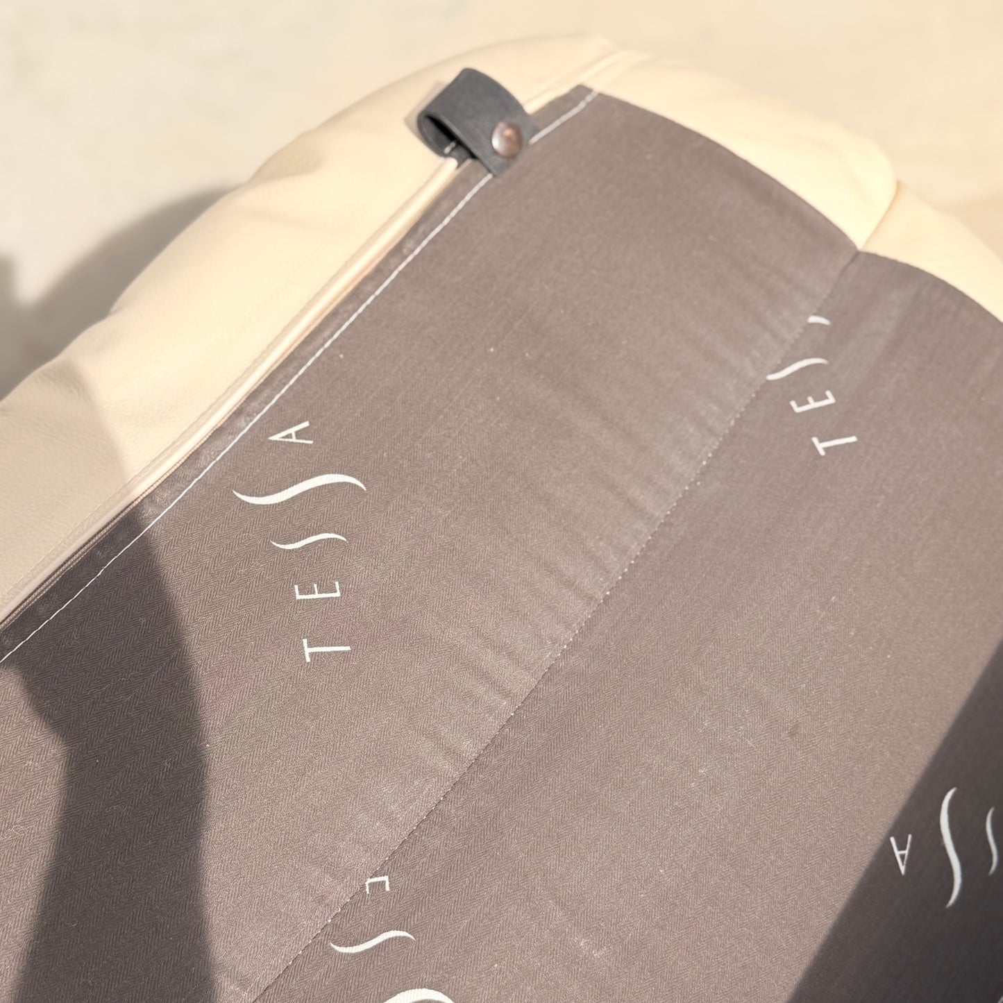Close-up of a beige and gray outdoor cushion with "TESSA" in white letters. Sunlight casts a shadow, complementing the look of the nearby Tessa Austin Blonde Leather Armchair (Revive).