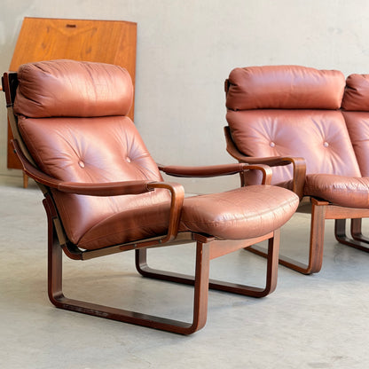 A modular two-seater sofa and armchairs from the Tessa T8 Lounge Suite (Revive) feature brown leather cushions and curved wood frames on a light floor, with a wooden cabinet in the background.