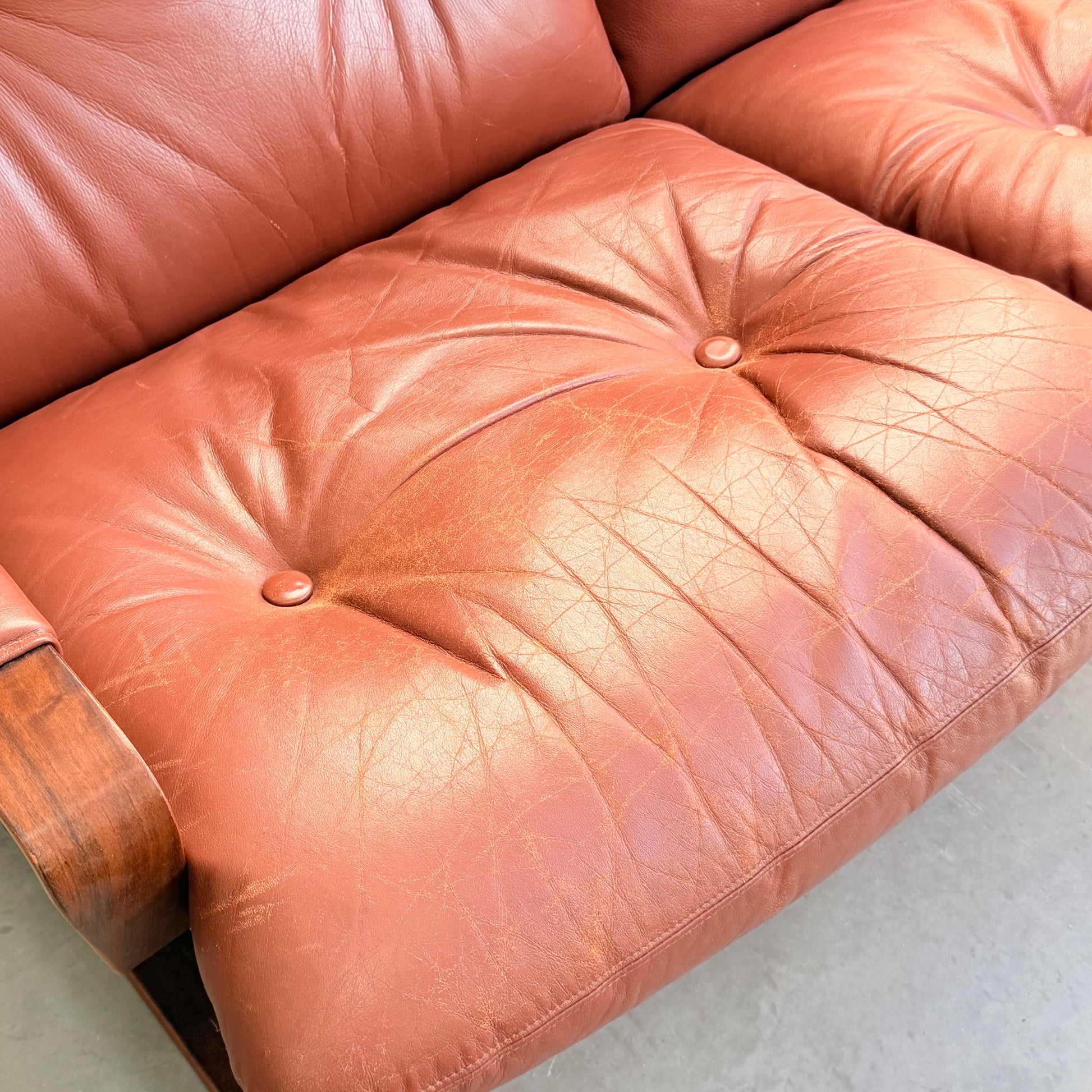 Close-up of a reddish-brown leather cushion from the Tessa T8 Lounge Suite modular two-seater sofa & armchairs (Revive), with visible creases, wrinkles, two tufted buttons, and part of a wooden armrest on the left.