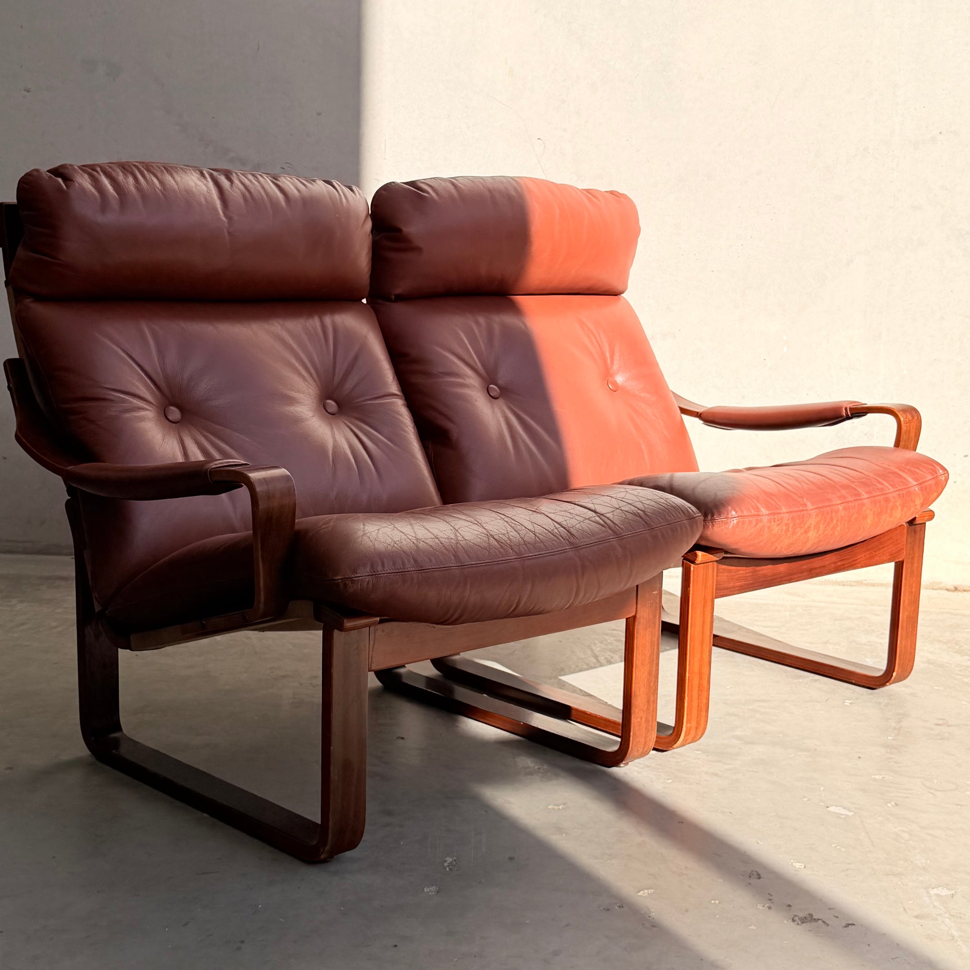 Two Tessa T8 Lounge Suite modular two-seater sofas and matching armchairs (Revive) with brown leather and wooden frames sit side by side in sunlight, casting shadows on a light-colored floor and wall.