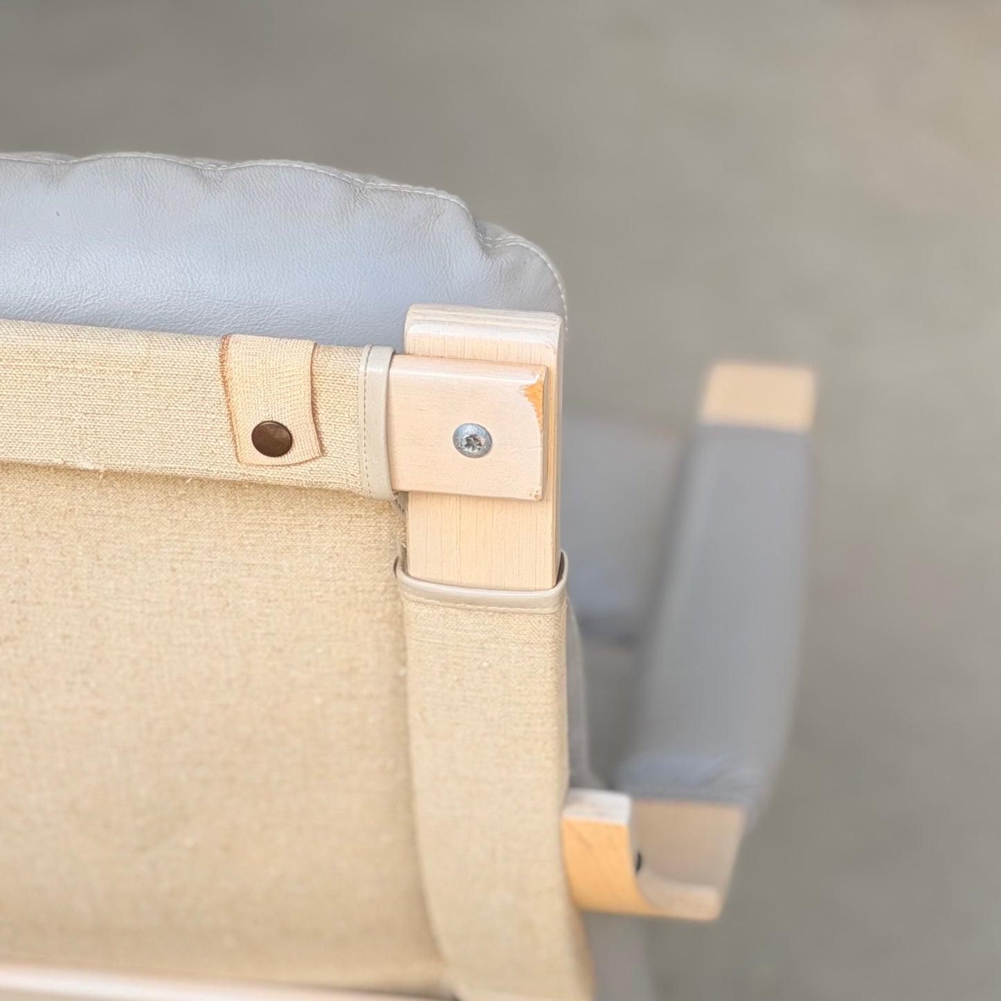 Close-up of the top corner of a Pair of Tessa Ferrara Swivel Armchairs (Revive), featuring light wood frames, beige canvas backrests, and grey cushioned headrests—a stunning example of mid-century Australian design. Blurred background.
