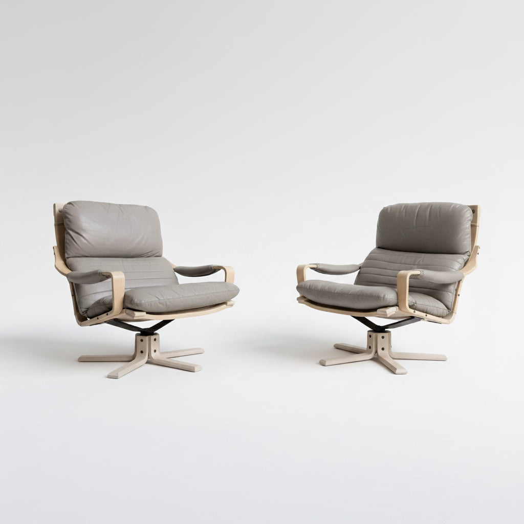 A pair of Tessa Ferrara Swivel Armchairs (Revive), featuring light wooden frames and grey cushioned seats, are positioned side by side against a plain light wall on a concrete floor.