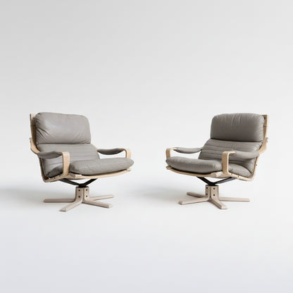 A pair of Tessa Ferrara Swivel Armchairs (Revive), featuring light wooden frames and grey cushioned seats, are positioned side by side against a plain light wall on a concrete floor.