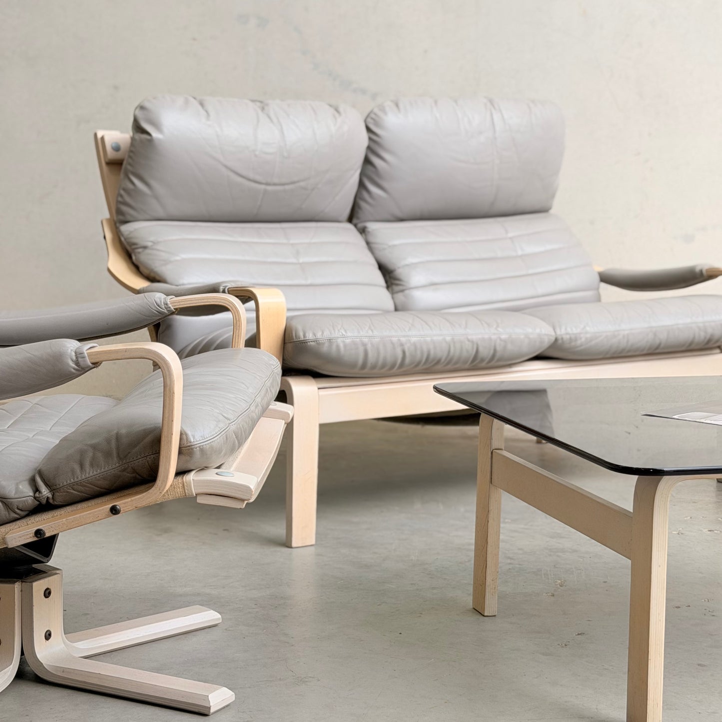 A modern living room showcases the Tessa Ferrara Lounge Suite – Complete 5-Piece Set (Revive) with gray cushions and light wood accents, arranged beside a glass-top coffee table on a light gray floor.