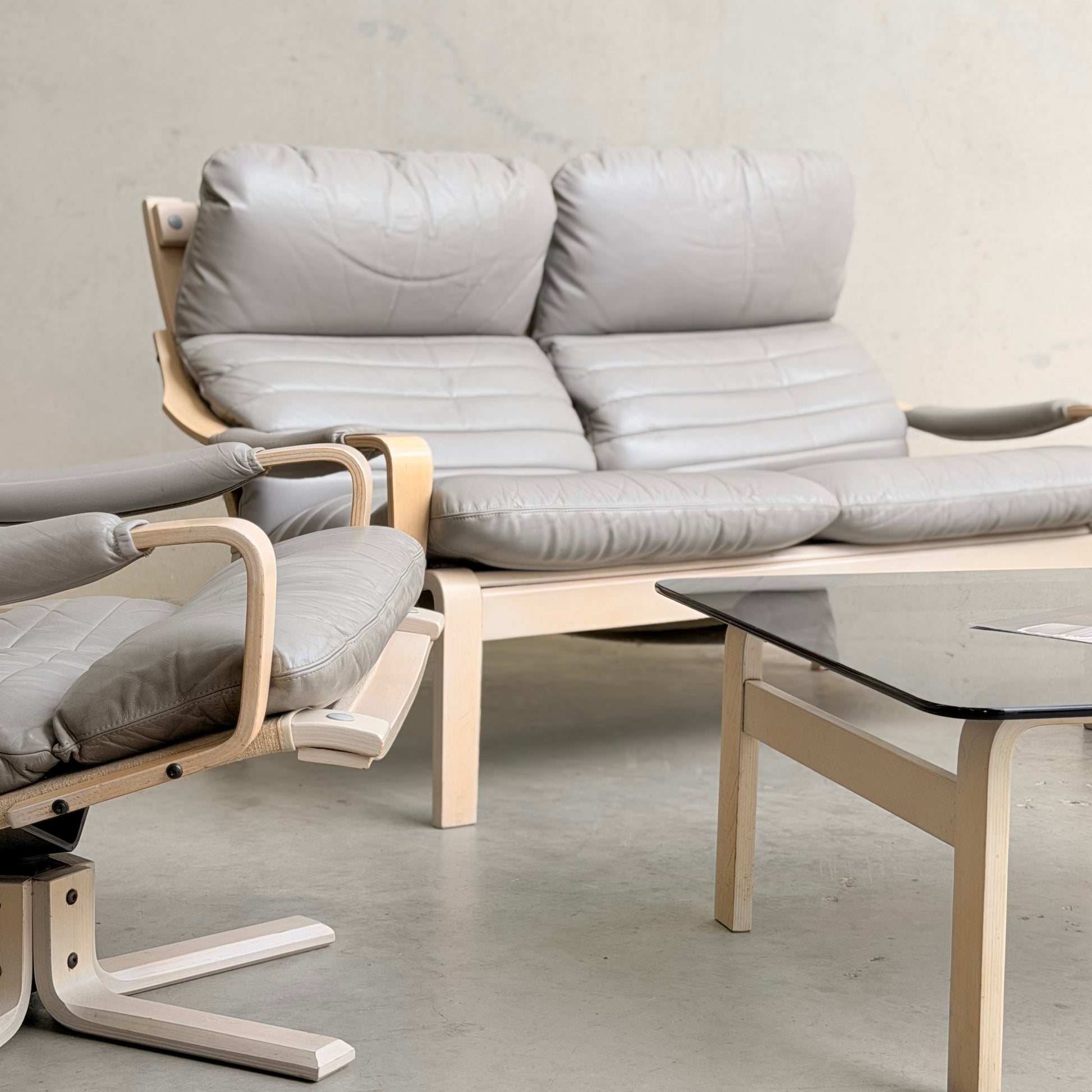 A modern living room showcases the Tessa Ferrara Lounge Suite – Complete 5-Piece Set (Revive) with gray cushions and light wood accents, arranged beside a glass-top coffee table on a light gray floor.