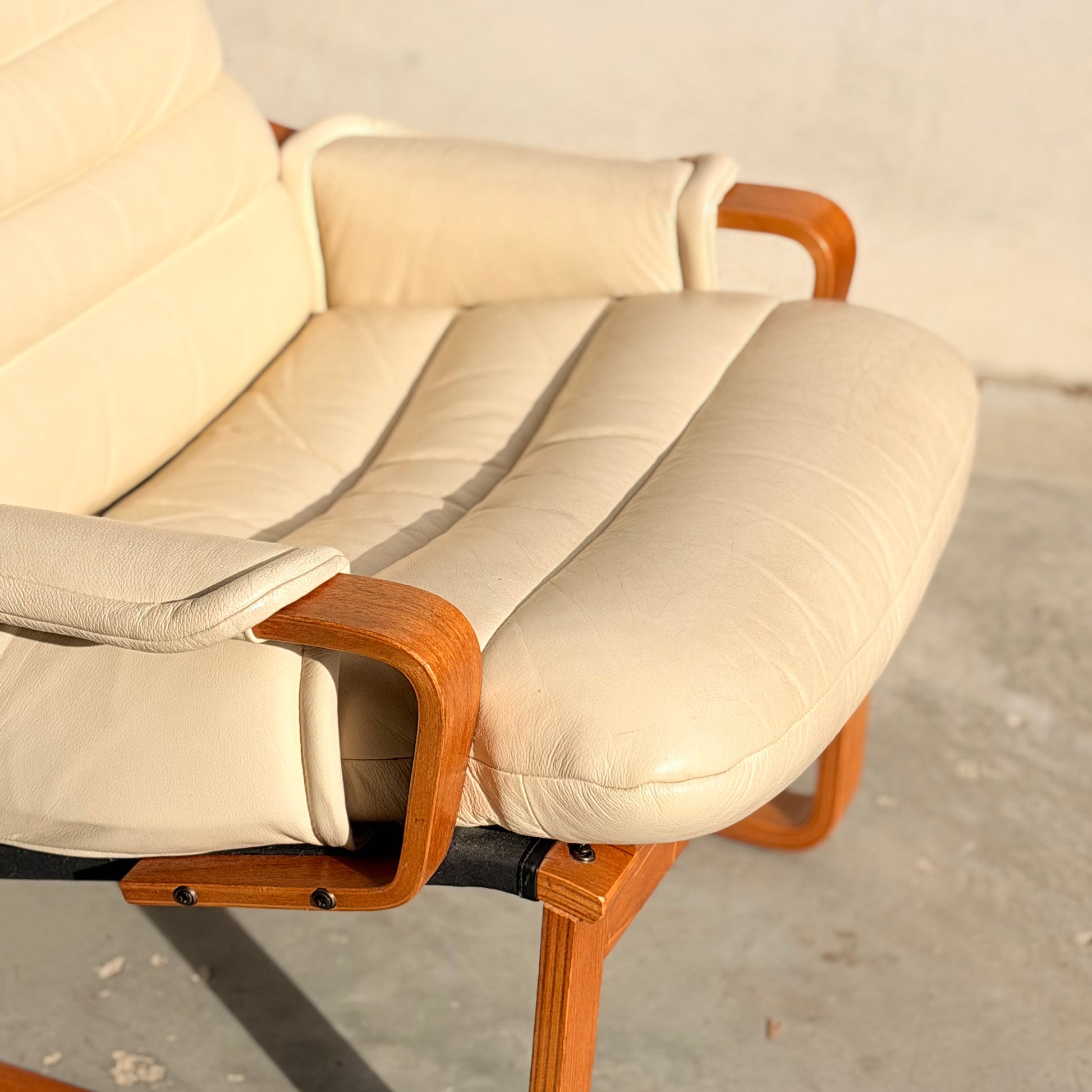 A close-up of the Tessa Austin Blonde Leather Armchair (Revive) with wooden armrests, positioned on a concrete floor against a light wall. Sunlight casts soft shadows on the beige leather.