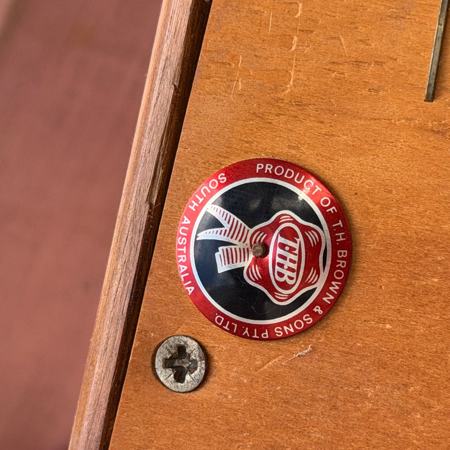 A round metal badge on wood, featuring a red and white helmet logo with "Product of F.H. Brown & Sons Pty Ltd South Australia" around the edge—an iconic detail on the Set of 3 Vintage THB Bar Stools - COMING SOON. A screw is visible nearby.