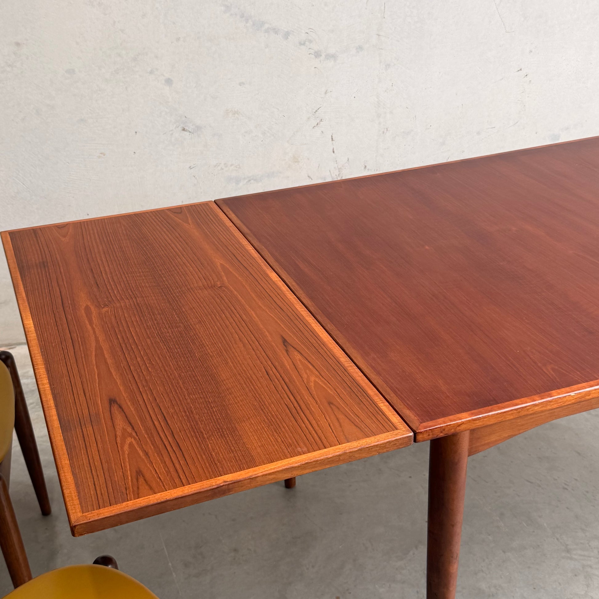 A Vintage Mid-Century Parker 109 Nordic Extension Dining Table (Revive) displays a subtle wood grain and color variation between the main teak surface and the extension, with two yellow chairs partly visible underneath.