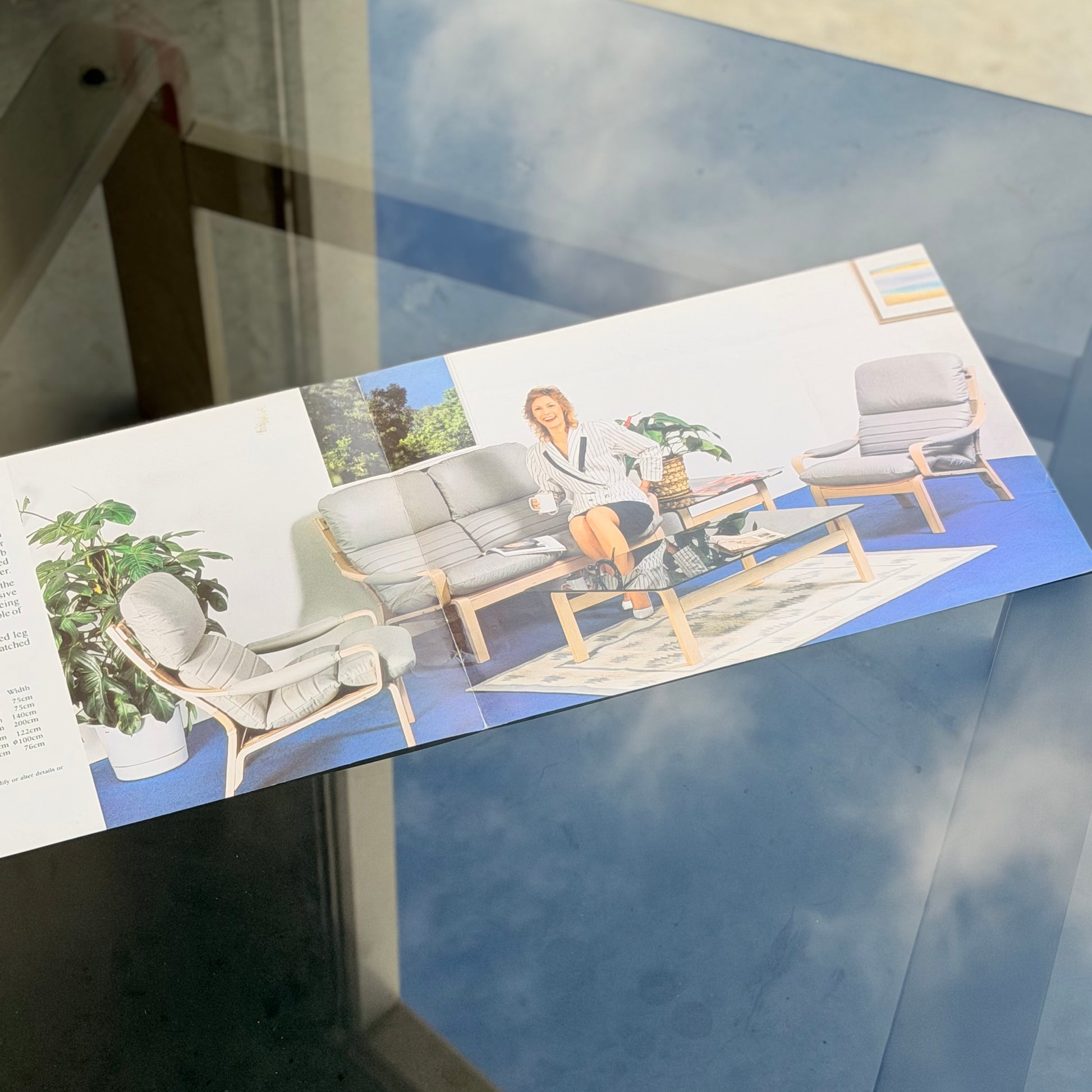 A brochure on a glass table shows a mid-century modern living room with gray furniture, including the Tessa Ferrara Lounge Suite – Complete 5-Piece Set (Revive), a woman seated on the sofa, potted plants, and artwork. Reflections of sky shimmer on the glass.