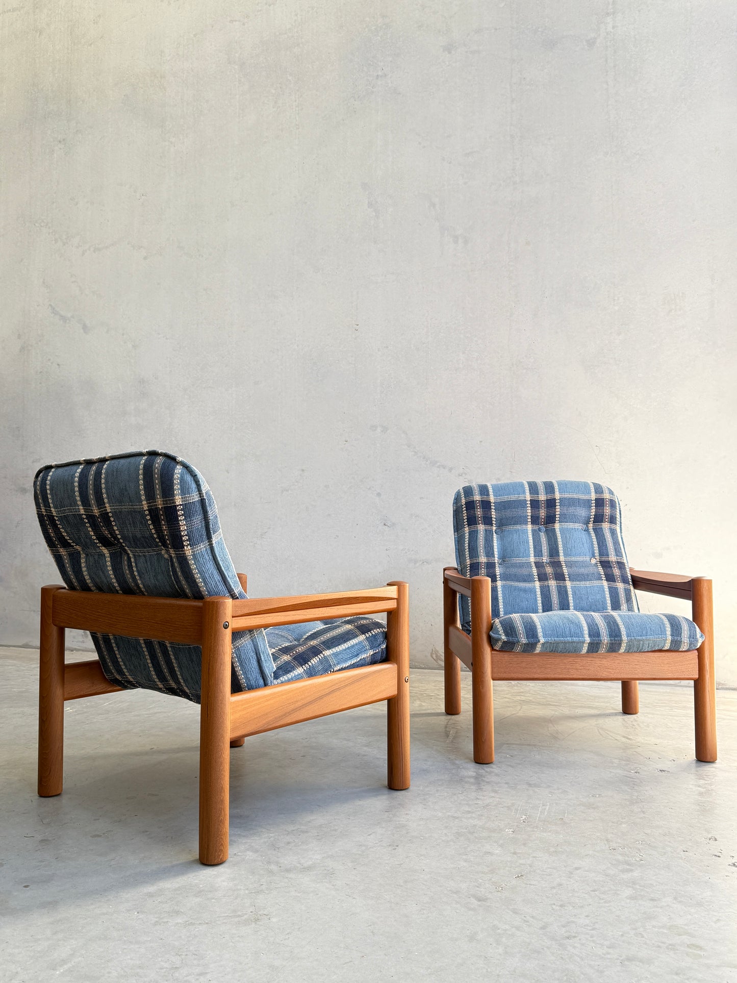 The Restored Domino Møbler Armchairs – Blue Plaid Mid-Century Set (Rediscover) features two Scandinavian solid wood chairs with retro blue plaid cushions, placed on a light concrete floor against a textured, light gray wall.
