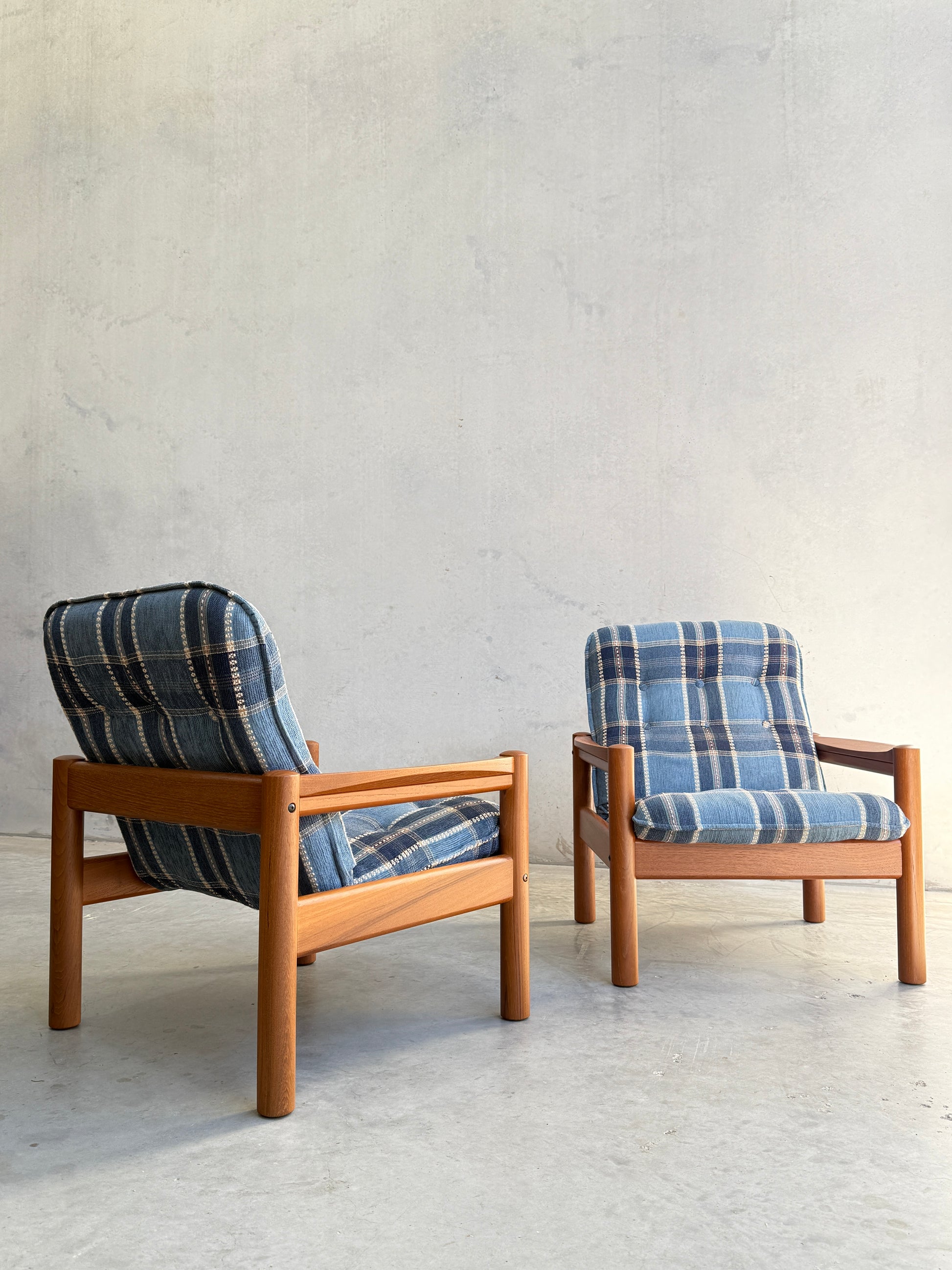 The Restored Domino Møbler Armchairs – Blue Plaid Mid-Century Set (Rediscover) features two Scandinavian solid wood chairs with retro blue plaid cushions, placed on a light concrete floor against a textured, light gray wall.