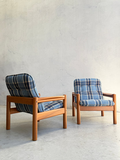 The Restored Domino Møbler Armchairs – Blue Plaid Mid-Century Set (Rediscover) features two Scandinavian solid wood chairs with retro blue plaid cushions, placed on a light concrete floor against a textured, light gray wall.