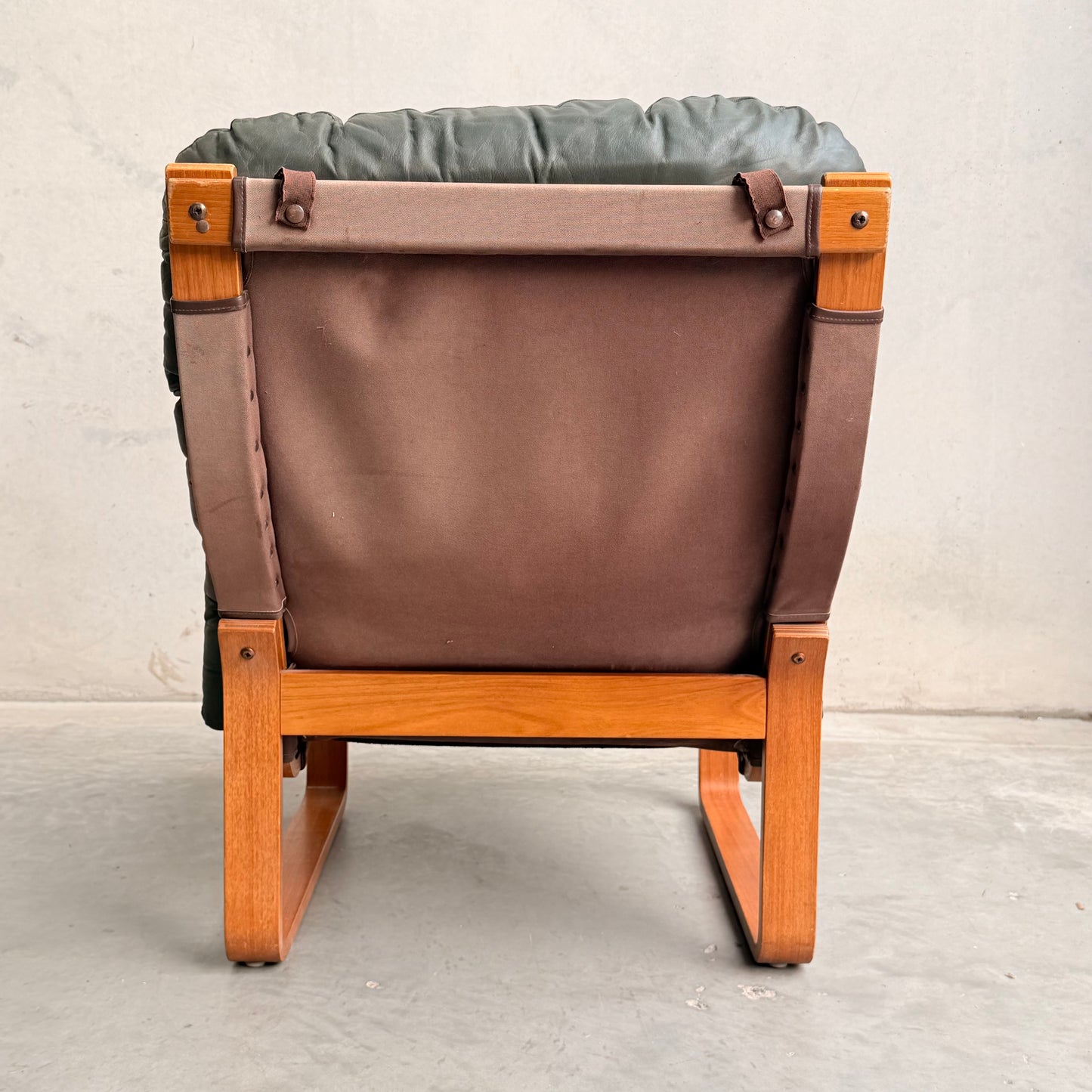The Tessa Vintage Single No Arm Monaco Armchair in Forest Green (Revive) features a wooden frame, brown fabric back, and dark green cushioned seat with leather accents, placed on a gray concrete floor against a light gray wall.