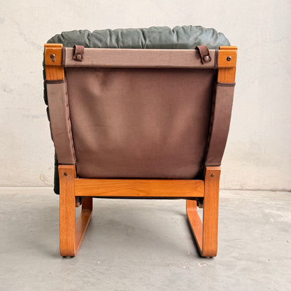 The Tessa Vintage Single No Arm Monaco Armchair in Forest Green (Revive) features a wooden frame, brown fabric back, and dark green cushioned seat with leather accents, placed on a gray concrete floor against a light gray wall.