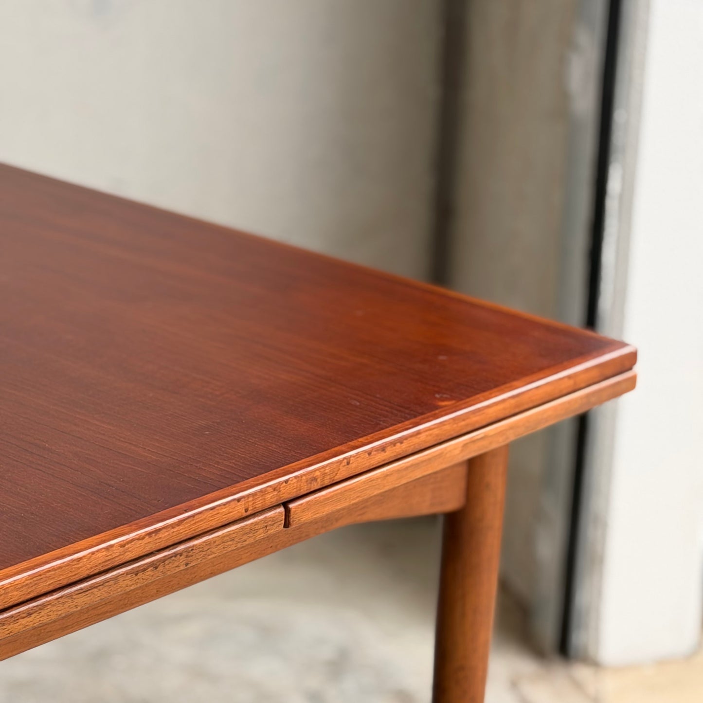 Close-up of the corner of a Vintage Mid-Century Parker 109 Nordic Extension Dining Table - Table Only (Revive), featuring smooth edges, a rich reddish-brown finish, and minimalist design near a concrete wall and partially open door.