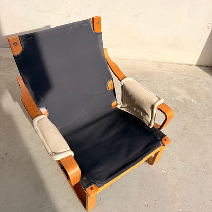 The Tessa Austin Blonde Leather Armchair (Revive) features a wooden frame, padded light armrests, and a black seat, set on a concrete floor in bright sunlight against a pale wall.