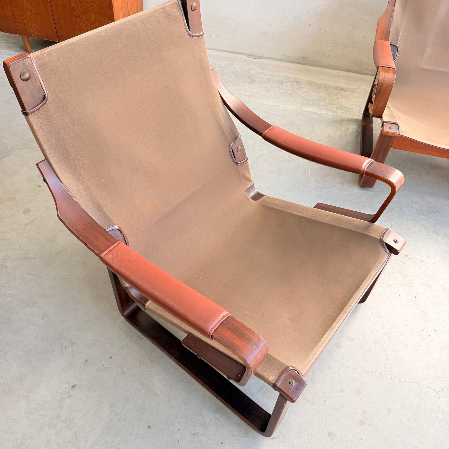 A modern lounge chair with brown canvas seat, wooden frame, and leather armrests sits on a concrete floor beside a matching Tessa T8 Lounge Suite Revive modular two-seater sofa and armchairs by Fred Lowen.