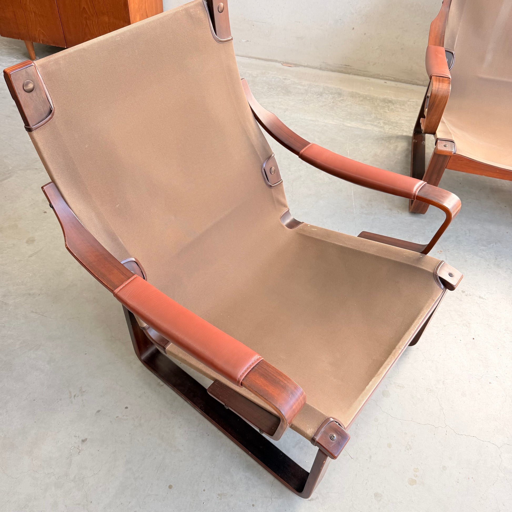 A modern lounge chair with brown canvas seat, wooden frame, and leather armrests sits on a concrete floor beside a matching Tessa T8 Lounge Suite Revive modular two-seater sofa and armchairs by Fred Lowen.