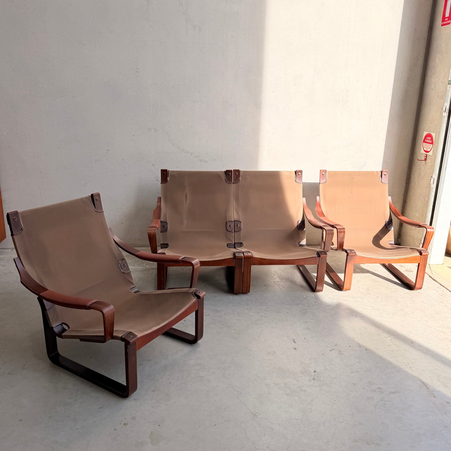 Three brown leather sling chairs, including a Tessa T8 Lounge Suite modular two-seater sofa and armchairs (Revive), sit in sunlight by a plain light wall on concrete, highlighting Fred Lowen's mid-century modern wooden frame design.