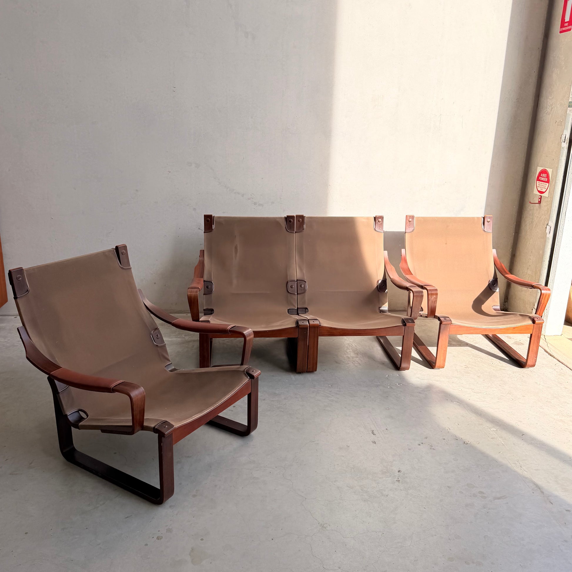 Three brown leather sling chairs, including a Tessa T8 Lounge Suite modular two-seater sofa and armchairs (Revive), sit in sunlight by a plain light wall on concrete, highlighting Fred Lowen's mid-century modern wooden frame design.