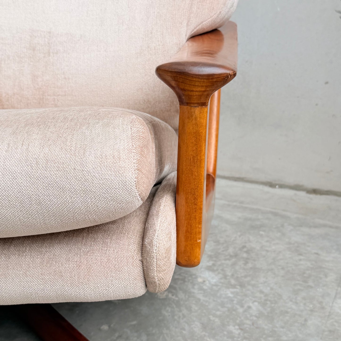 Close-up of the Vintage Mid Century Tessa T21 Swivel Chair (Revive) featuring beige upholstery and a curved wooden armrest and frame, set on a light gray concrete floor against a plain wall.
