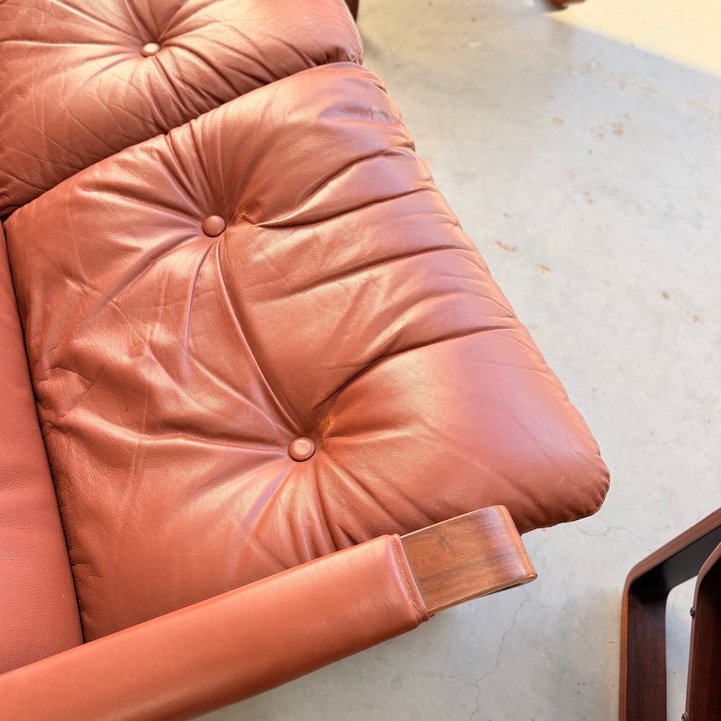 Close-up of a tufted, brown leather cushion on the Tessa T8 Lounge Suite modular two-seater sofa & armchairs (Revive), mid-century modern style with wooden armrests, set on a light-colored concrete floor.