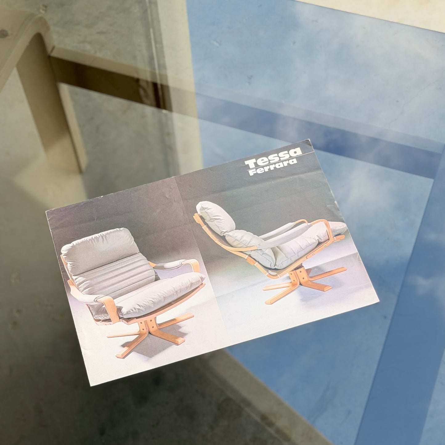 A brochure featuring the Tessa Ferrara Lounge Suite – Complete 5-Piece Set (Revive), two wooden chairs with gray cushions, sits on a glass table reflecting the blue sky—evoking timeless mid-century modern style.