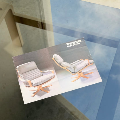 A brochure featuring the Tessa Ferrara Lounge Suite – Complete 5-Piece Set (Revive), two wooden chairs with gray cushions, sits on a glass table reflecting the blue sky—evoking timeless mid-century modern style.