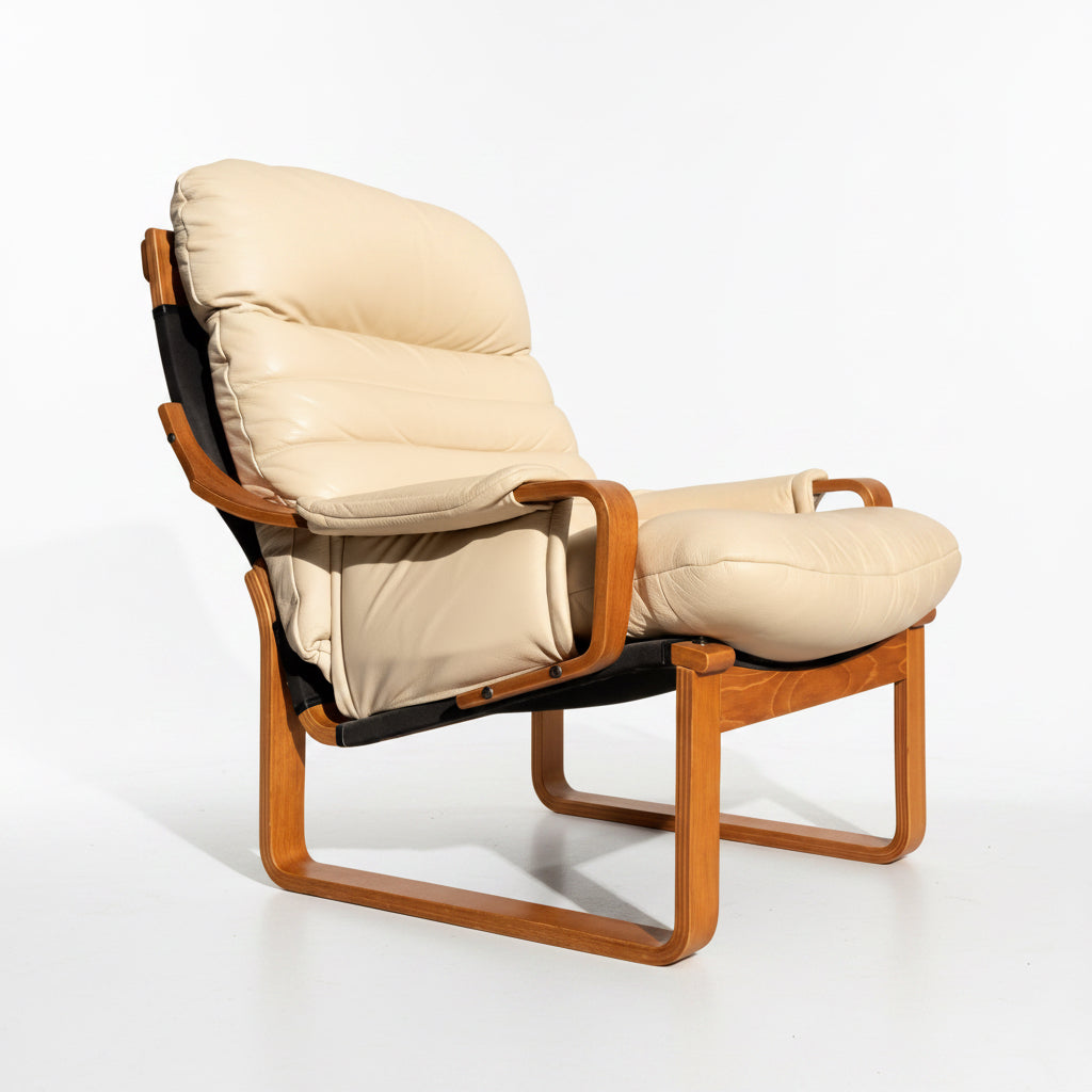 The Tessa Austin Blonde Leather Armchair (Revive) features plush beige leather cushions and a curved wooden frame, set on a concrete floor against a plain light-colored wall.
