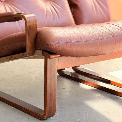Close-up of the Tessa T8 Lounge Suite two-seater sofa and armchairs (Revive), featuring vintage brown leather, wooden armrests and legs, with textured, slightly worn upholstery in natural light on a concrete floor.