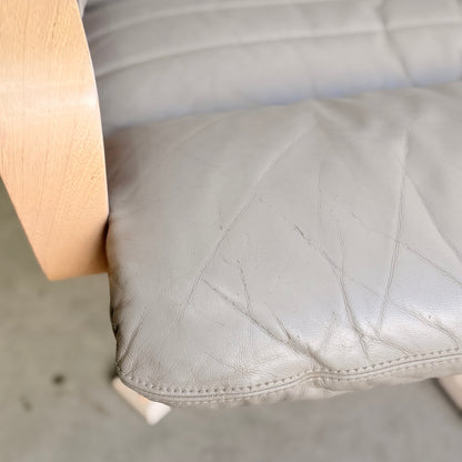Close-up of a light gray, gently wrinkled leather chair cushion with a light wooden armrest on the left. The mid-century style mirrors the Tessa Ferrara Lounge Suite – Complete 5-Piece Set (Revive). The background is blurred.