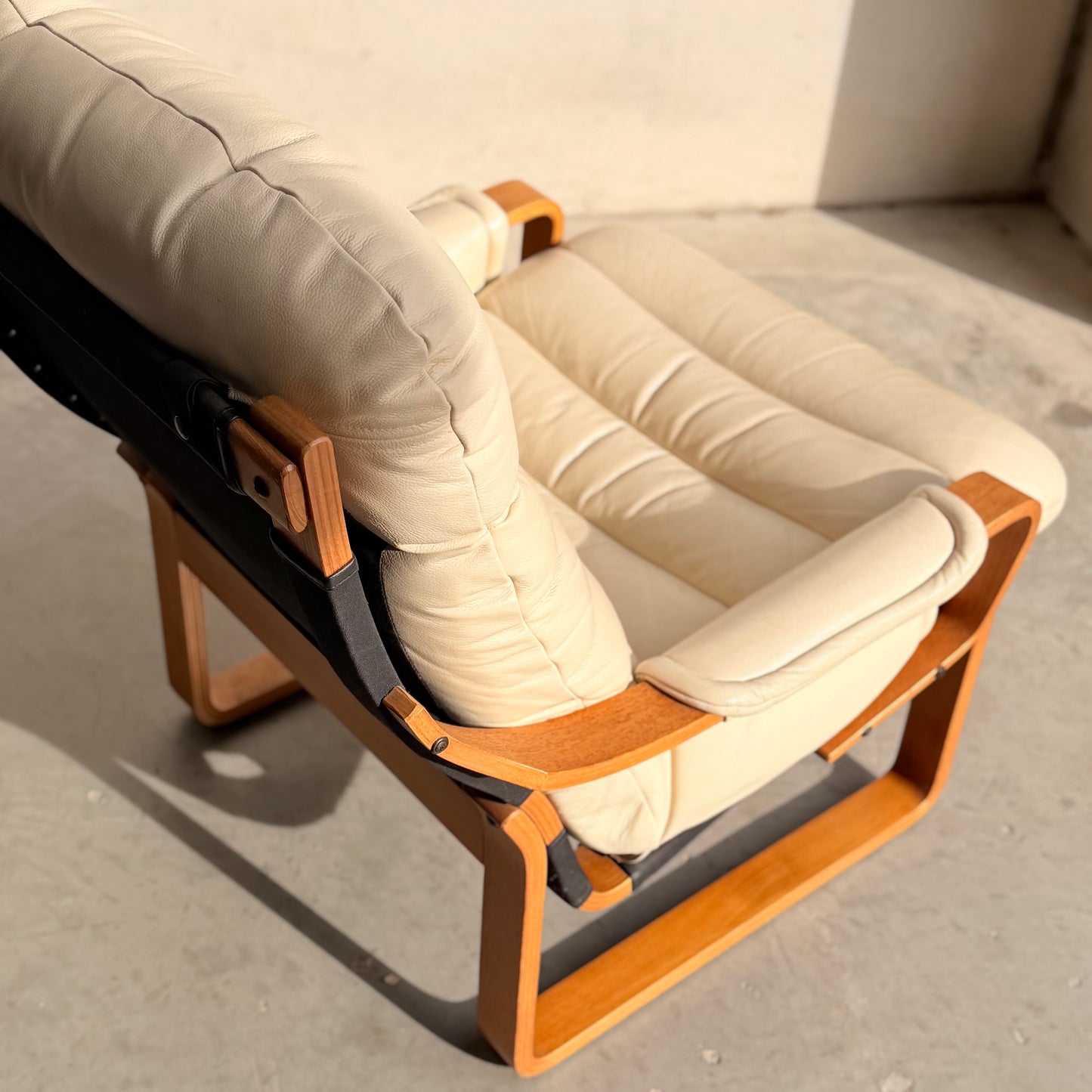 The Tessa Austin Blonde Leather Armchair (Revive), featuring light beige leather cushions and a wooden frame, sits on a concrete floor bathed in warm sunlight.