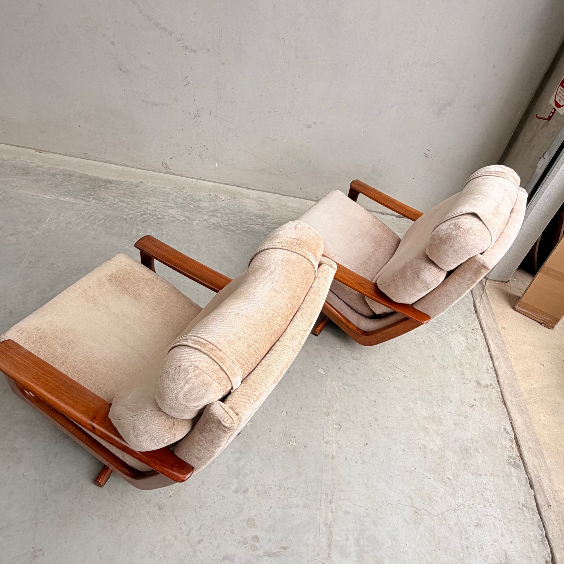 Two Vintage Mid Century Tessa T21 Swivel Chairs (Revive) with beige cushions and wooden frames sit side by side at a slight angle on a gray concrete floor against a plain, light-colored wall.