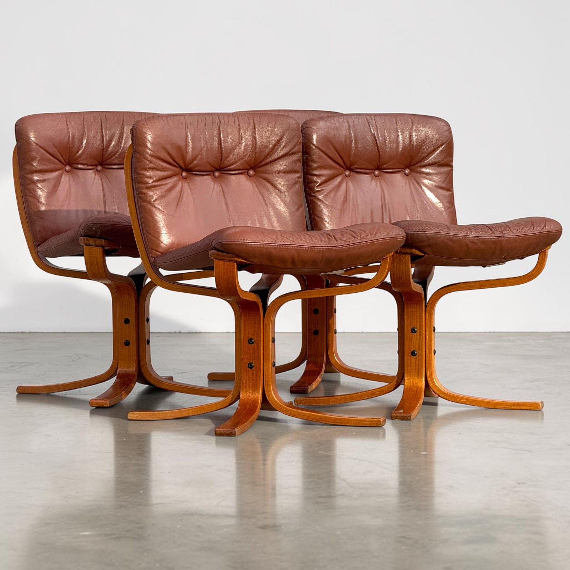 A rare set of 4 vintage Joe Rufenacht lounge chairs in Howe leather, featuring curved wooden frames, are grouped on a polished concrete floor, exuding Scandinavian style against a plain light background.