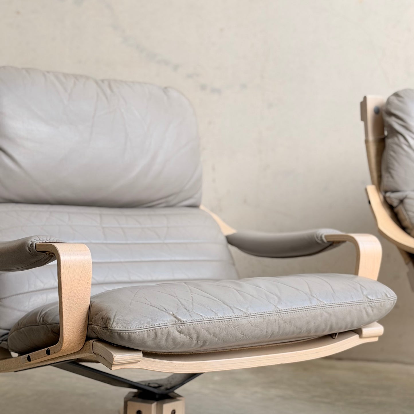 Close-up of a Pair of Tessa Ferrara Swivel Armchairs (Revive) with light beige leather cushions and a light wood frame, set against a textured wall, reflecting mid-century Australian design. Another chair is partially visible in the background.