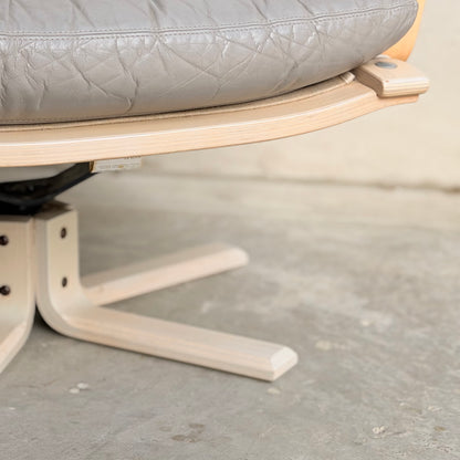 Close-up of the base and seat of a Pair of Tessa Ferrara Swivel Armchairs (Revive) with light wood legs and quilted gray leather upholstery, set on a concrete floor near a beige wall.