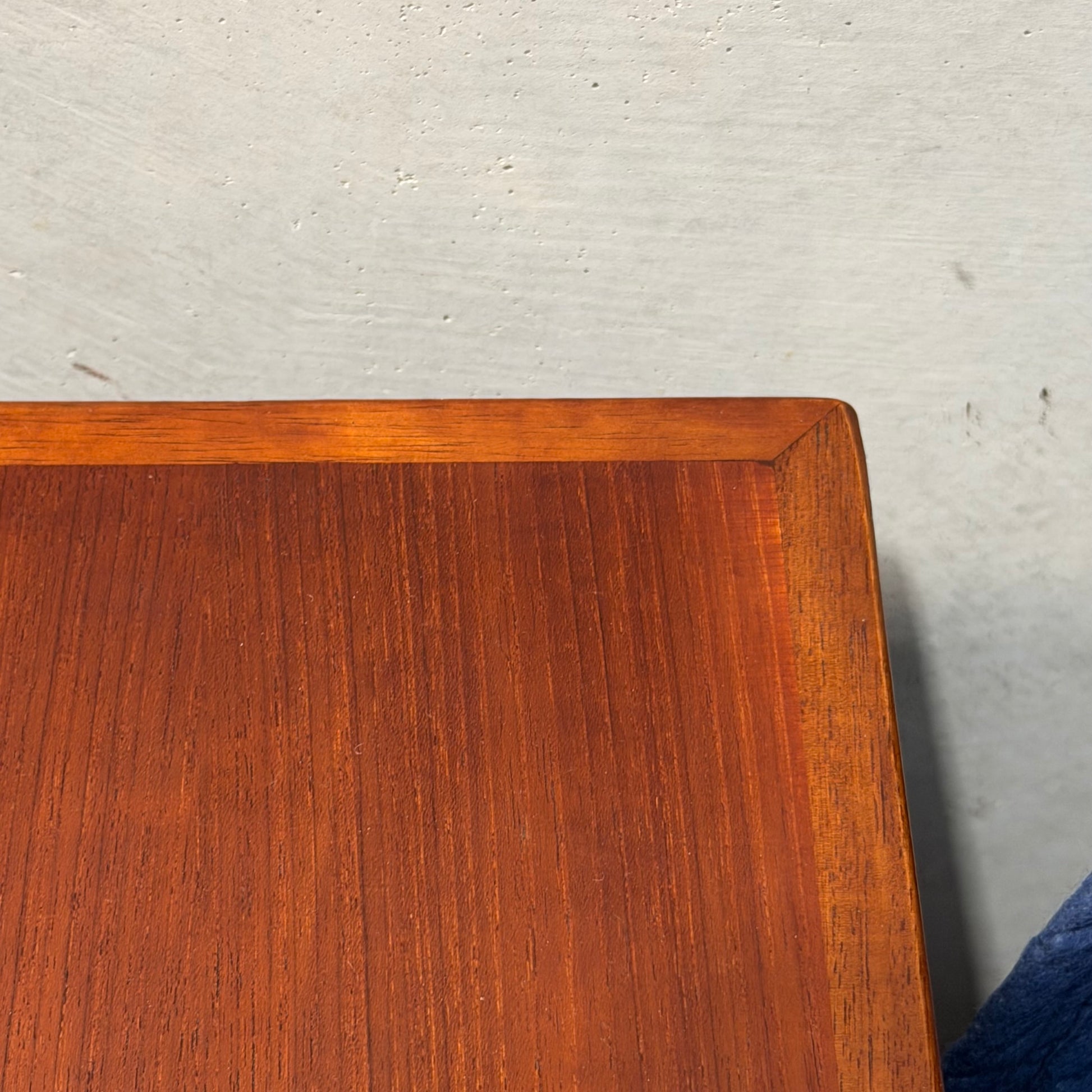 Close-up of the corner of a Vintage Mid-Century Parker 109 Nordic Extension Dining Table (Revive) with visible teak grain and reddish-brown finish, set against a light gray textured wall. A hint of a dark blue object appears at the edge.