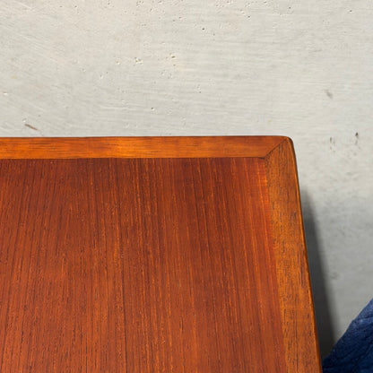 Close-up of the corner of a Vintage Mid-Century Parker 109 Nordic Extension Dining Table (Revive) with visible teak grain and reddish-brown finish, set against a light gray textured wall. A hint of a dark blue object appears at the edge.