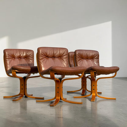 A rare set of 4 vintage Joe Rufenacht lounge chairs in Howe leather, featuring tufted backs and wooden frames, are arranged in a sunlit room with a gray floor and plain white wall.