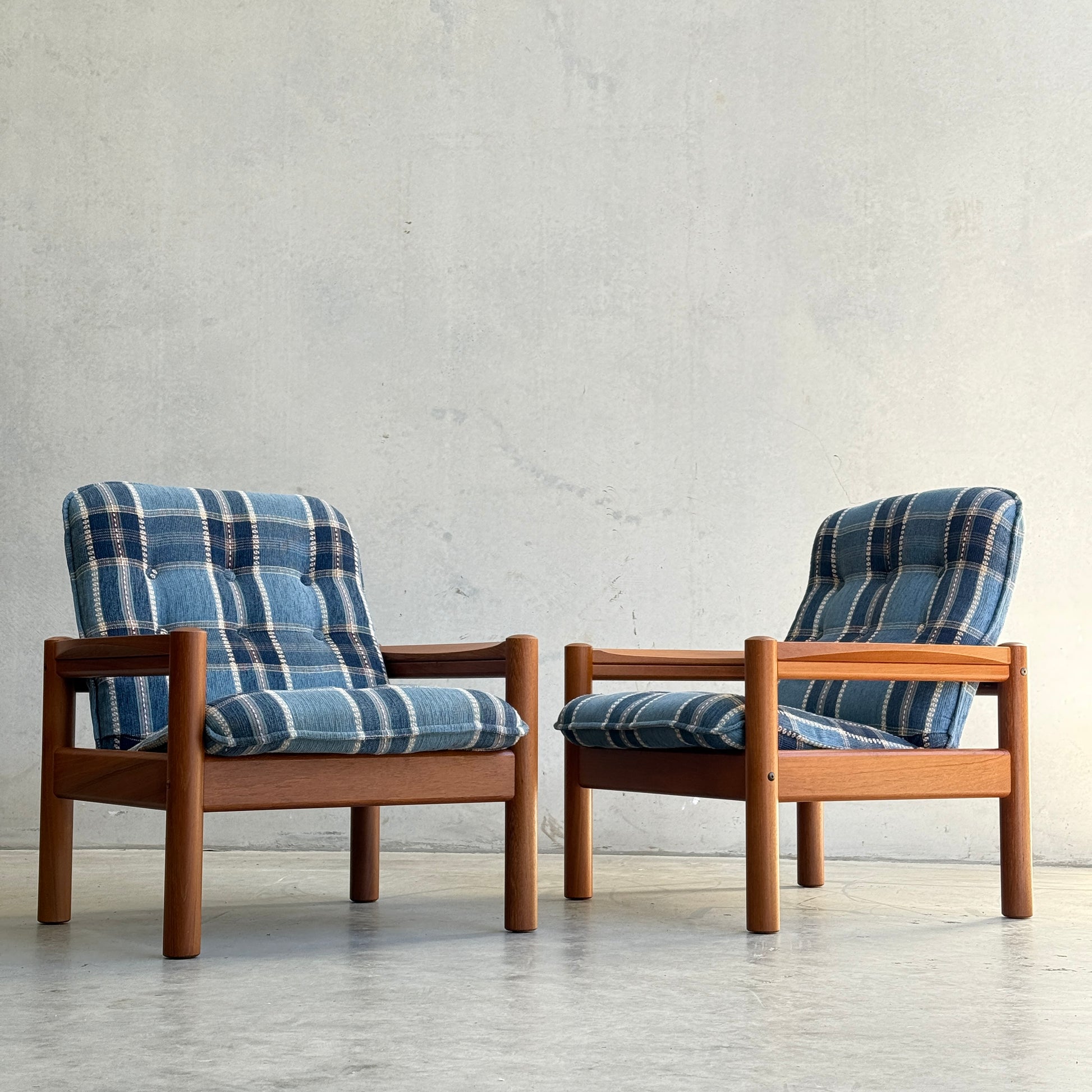 A pair of Restored Domino Møbler Armchairs – Blue Plaid Mid-Century Set (Rediscover) stand side by side on a concrete floor, their solid wood frames and retro plaid cushions set against a plain light wall.